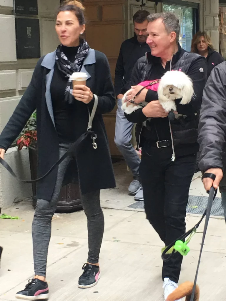Two people walk on a city sidewalk; one holds a coffee and a dog leash, the other carries a small white dog dressed in pink. Other people and a dog are visible in the background.
