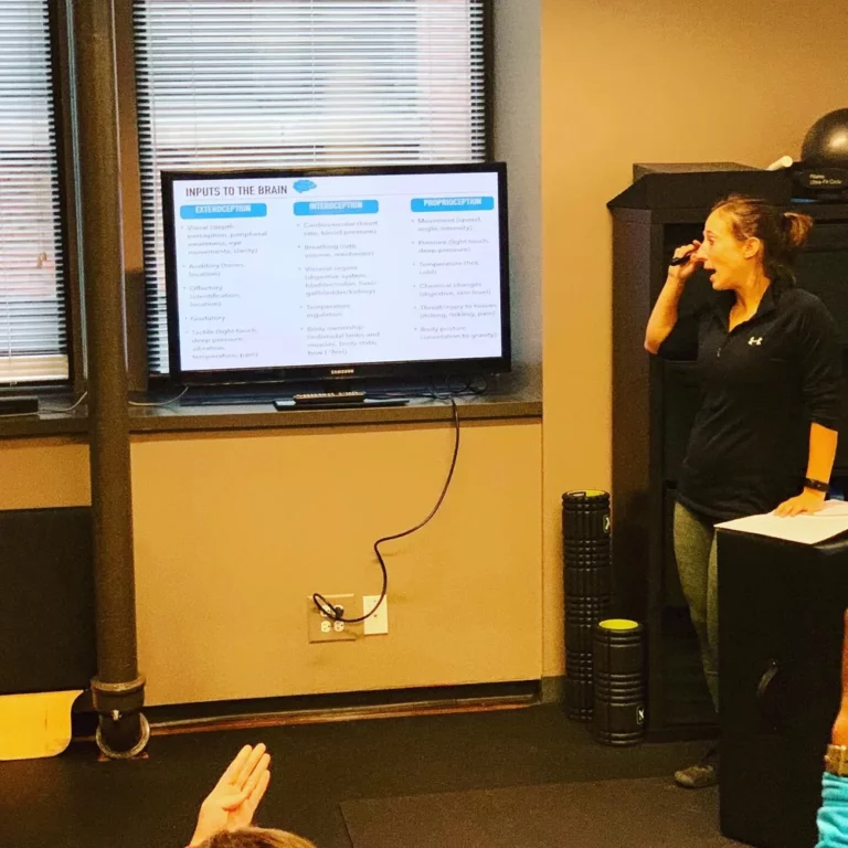 A woman stands beside a screen displaying a presentation titled "INPUTS TO THE BRAIN" while speaking to an audience in a room.
