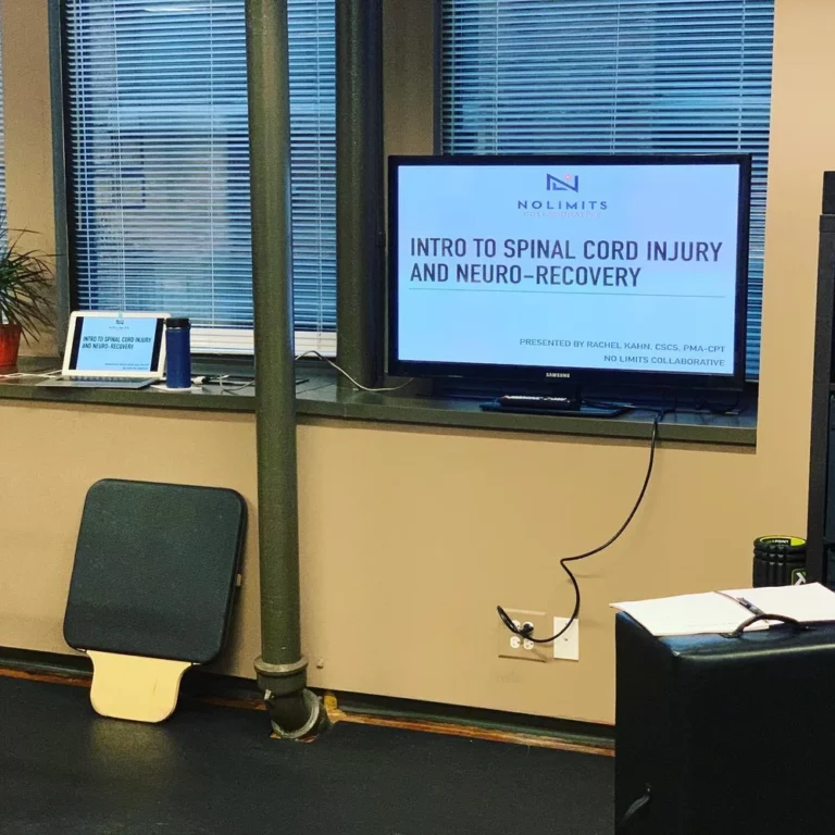 A TV screen displays a presentation titled "Intro to Spinal Cord Injury and Neuro-Recovery" in an office setting with a window, plant, small sign, and exercise equipment.
