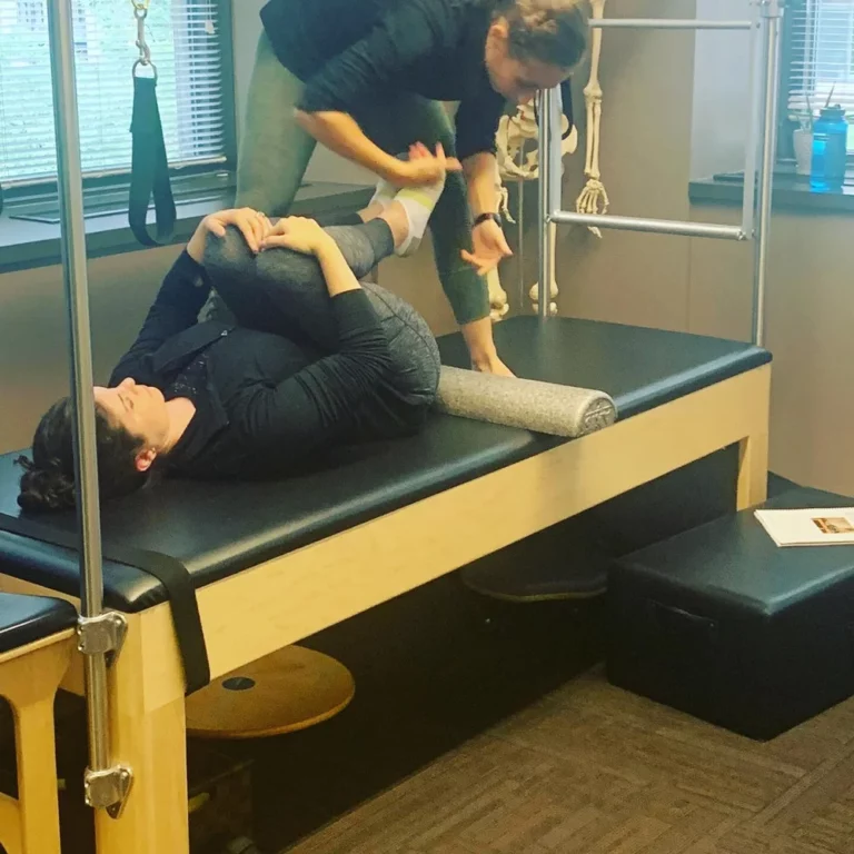 A woman lies on a Pilates reformer bench with her knees to her chest while an instructor guides her movement, in a fitness studio.