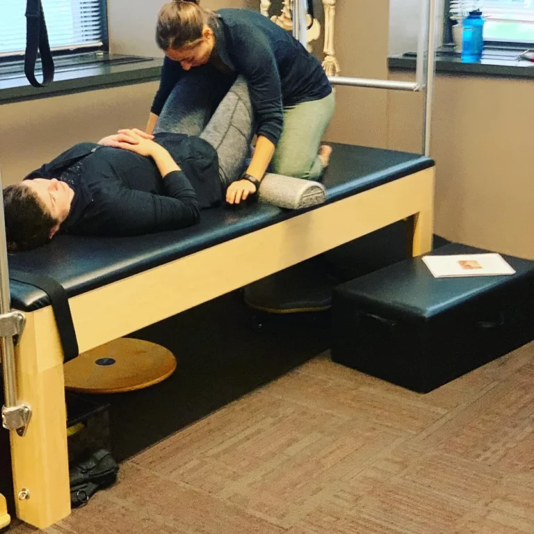 A physical therapist assists a patient lying on a padded table in a clinical setting, using a foam roller under the patient’s knee.