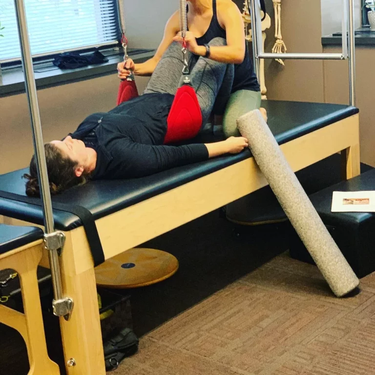 A person lies on a Pilates reformer with their legs elevated in straps, assisted by an instructor. A foam roller rests nearby on the platform.