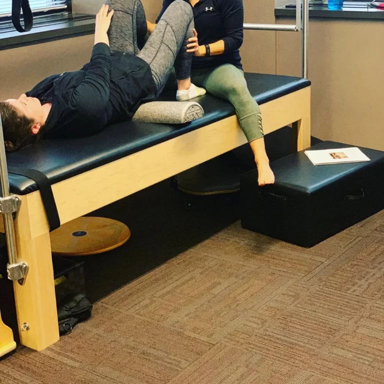 Two people in athletic wear are on a therapy table; one lies on their back with legs bent while the other assists, likely during a physical therapy session.