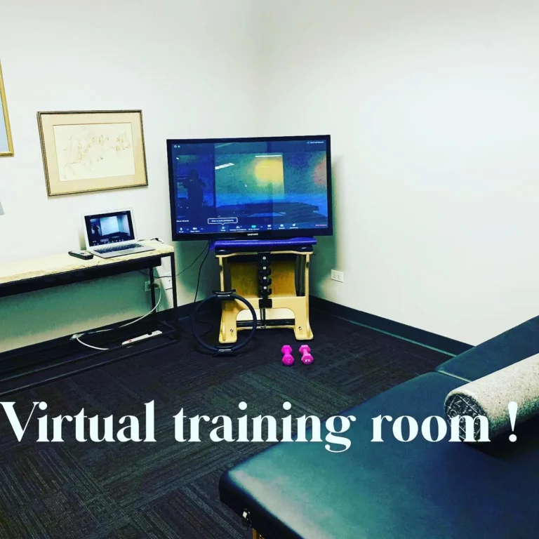 A small room with a massage table, a TV screen displaying a virtual session, a laptop on a side table, two pink dumbbells, and the text "Virtual training room!" on the floor.