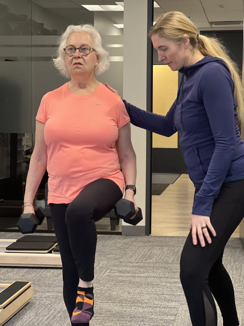 An older woman lifts one knee while holding dumbbells, guided by a trainer in a gym—a scene from Parkinson’s Exercise Programs in Chicago designed to support strength and mobility.