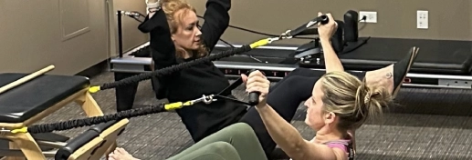 Two women exercise on Pilates reformer machines, pulling resistance bands with both hands during small group fitness classes in Chicago’s modern fitness studio.